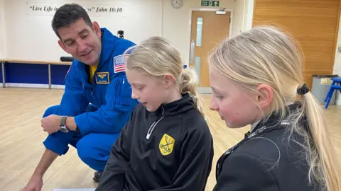 SAM READ/BBC Commander Jack Hathaway is wearing blue NASA overalls and kneeling down in a school hall talking to two young girls with blonde hair who are sitting on the floor.