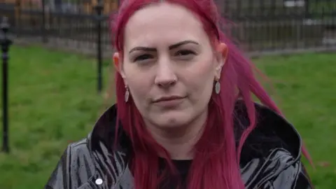 BBC A woman with red hair, staring at the camera, wearing a dark shiny coat