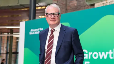 West Midlands Combined Authority West Midlands Mayor Richard Parker, wearing a navy blue suit and a white shirt with a red and white striped tie. He is standing in front of a green and blue branded board.