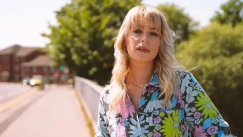 Rhiannon Holland Jess Davies wears a colourful floral boiler suit. She has wavy blonde hair with a fringe. She leans against a railing, with the road in the background blurred. 